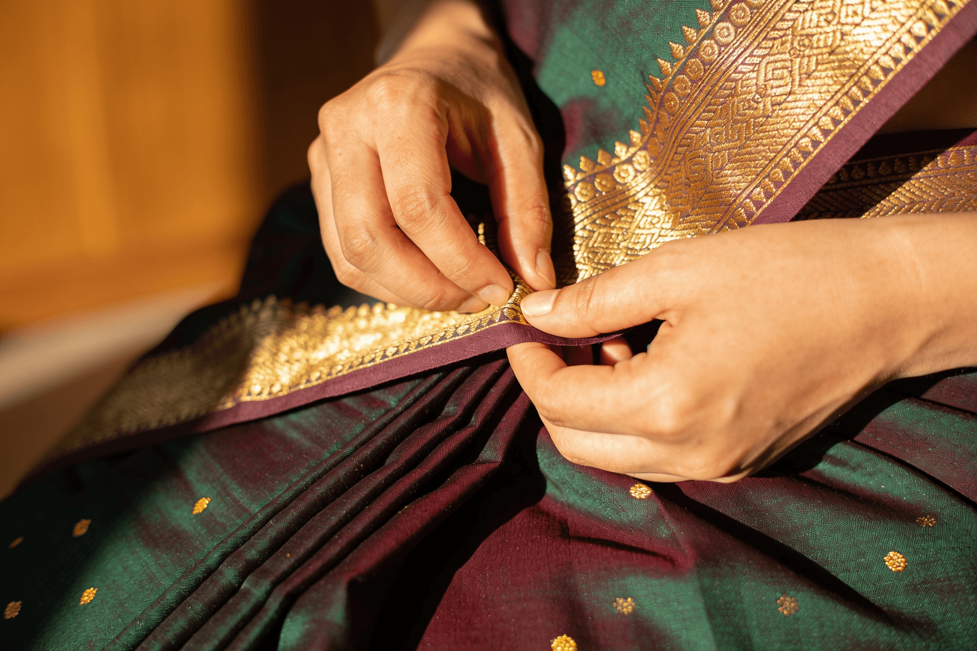 Close-up of hands pinning pleats on a Kanchipuram silk saree, showing the zari border detail