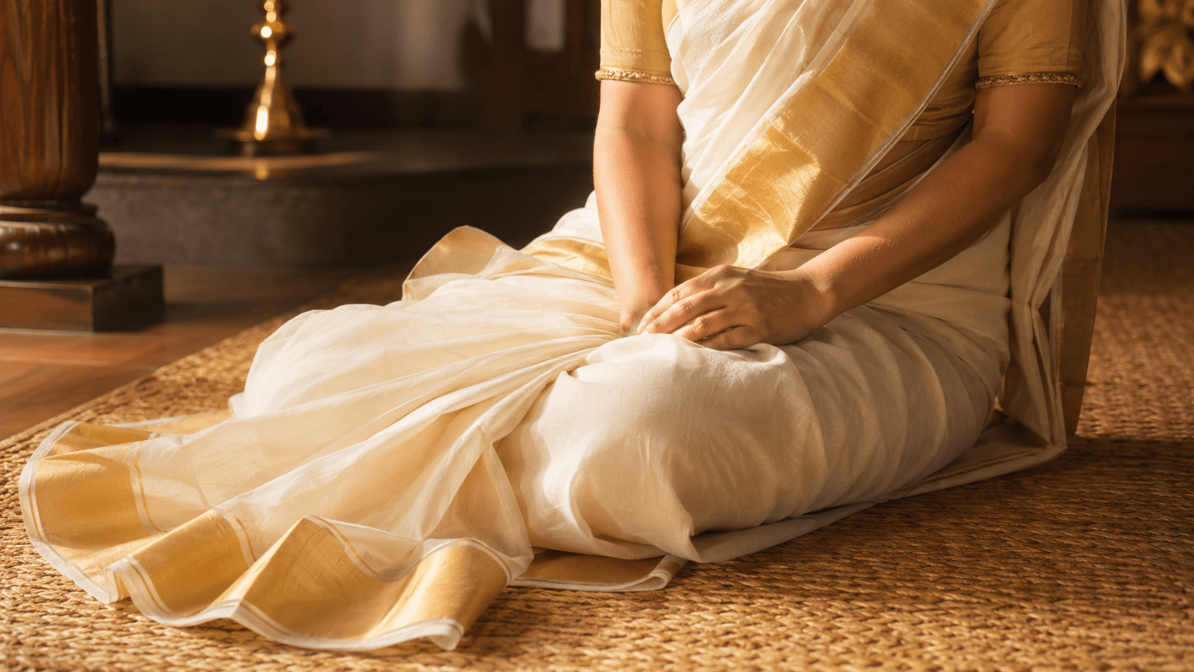 Woman in a Kerala kasavu saree seated on a woven mat on the floor, hands resting in her lap, gold zari border visible, warm natural light